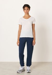 Biele tričko s véčkovým výstrihom v kombinácii s tmavomodrými teplákmi so stranovým logom. Tenisky sú sivé s bielymi akcentmi. Viditeľné sú hladké textúry látky.