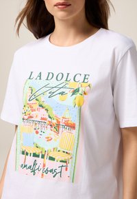 Hvid bomulds T-shirt med et farverigt grafisk tryk af en malerisk terrasse ved Amalfikysten, med et bord, stoler og citronakcenter.