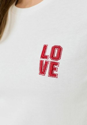 Biały materiał z czerwonym haftem liter układających się w słowo „LOVE” w dwurzędowym układzie na wysokości klatki piersiowej.