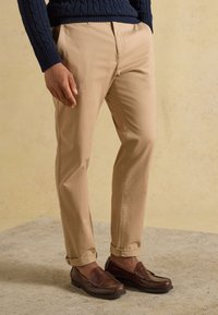 Homme portant un pantalon beige retroussé, des mocassins en cuir marron et un pull en maille torsadée bleu marine, debout sur un sol texturé.