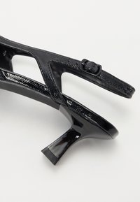 Černé lakované sandály s nízkým blokovým podpatkem, vybavené křížovými pásky a bočním uzávěrem na přezku. Hladká textura a elegantní design.
