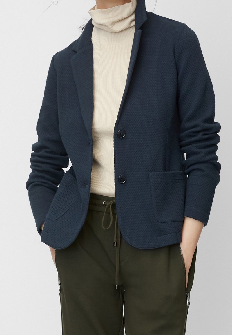 Blazer texturizado azul marino con solapa de muesca, dos bolsillos frontales y botones negros, usado sobre un cuello de tortuga crema y pantalones verde oliva.