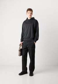 Felpa grigia con cappuccio con la scritta "targa" sul davanti, abbinata a jeans in denim scuro larghi e sneakers nere. Porta una borsa beige.