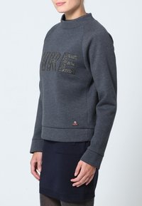 Donna che indossa una felpa grigia scura con la scritta "FIRE" sul petto e una gonna blu navy, in piedi contro uno sfondo chiaro e uniforme.