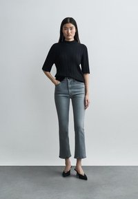 Žena stojaca, oblečená v čiernom svetri s krátkym rukávom, sivých džínsach s vysokým pásom a čiernych topánkach s ostrou špičkou, na jednoduchom sivom pozadí.