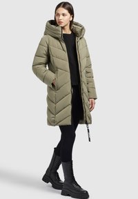 Abrigo acolchado largo color verde oliva con capucha, que presenta un acolchado en forma de chevrón, bolsillos laterales y cierre con cremallera. Se lleva con leggings negros y botas.