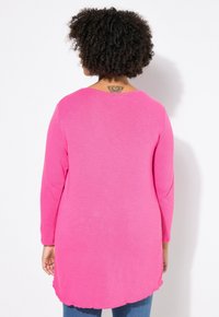 Langärmliges Oberteil aus leuchtend pinkem Stoff, runder Ausschnitt, gewellter Saum und Seitenschlitze. Von hinten gesehen, mit einem Tattoo am Nacken.