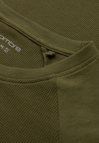 Oliivivihreä t-paita, jossa on teksturoitua, kuvioitua kangasta ja sileitä yksityiskohtia. Varustus pyöreällä kaula-aukolla ja näkyvillä ompeluyksityiskohdilla.