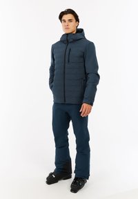Man in een donkerblauwe skijas met capuchon en bijpassende skibroek, staand met handen gedeeltelijk in de zakken, met zwarte skischoenen op een witte achtergrond.