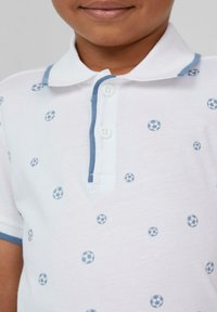 Polo blanc avec des accents bleu clair et de petits motifs de ballons de football. Il présente un col pointu et une patte de boutonnage à deux boutons. Texture douce.