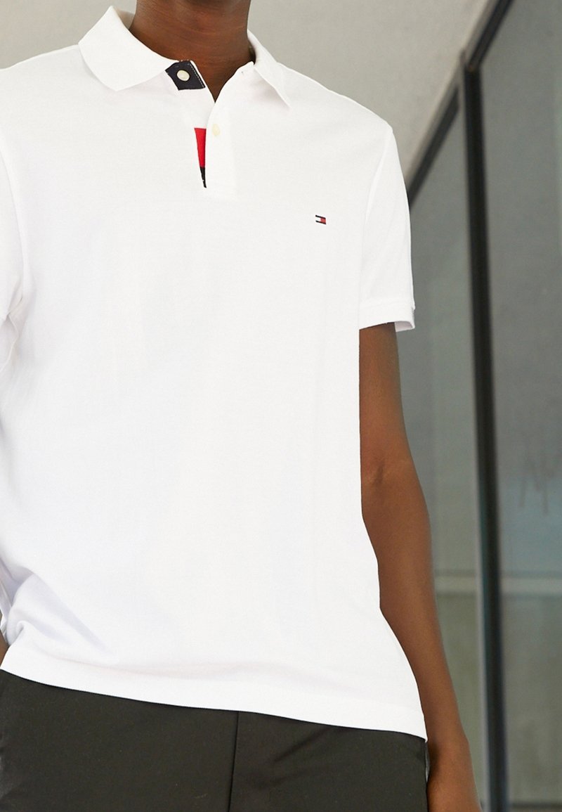 Personne portant un polo blanc à manches courtes avec un petit logo sur la poitrine et un pantalon noir, debout à l'intérieur près d'un mur en verre.