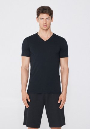 Camiseta negra de cuello en V, de manga corta, con un diseño ajustado. Tela suave y ligera. El modelo lleva pantalones cortos negros y está de pie contra un fondo neutro.
