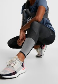 Černé a šedé sportovní legíny s různými texturami, přiléhavým střihem a vzorem z reflexních pruhů, doplněné o vícebarevné sportovní boty.