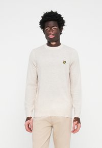 Pull en maille beige à manches longues et décolleté rond. Présente un petit logo en forme de papillon jaune sur le côté gauche de la poitrine. Texture douce.