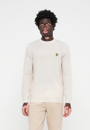 Pull en maille beige à manches longues et décolleté rond. Présente un petit logo en forme de papillon jaune sur le côté gauche de la poitrine. Texture douce.