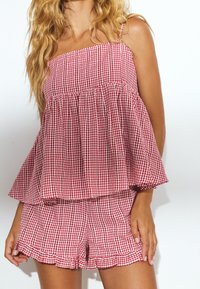Nainen, jolla on punavalkoinen ruudullinen hihaton toppi ja siihen sopivat röyhelöreunaiset shortsit valkoista taustaa vasten.