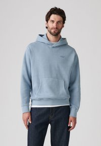 Lys blå hettegenser med en frontlomme, som har et enkelt design og en liten logo på brystet. Brukt over en hvit skjorte.