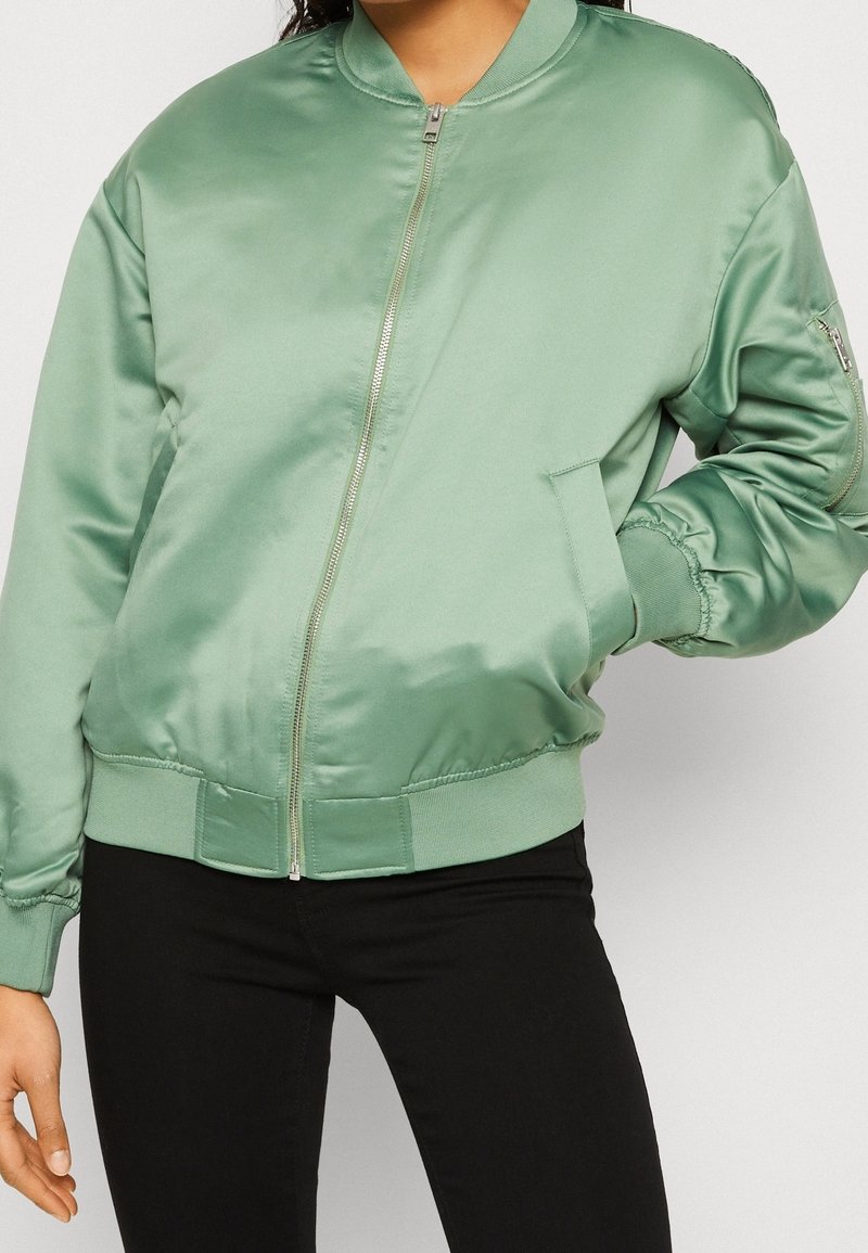 Femme portant une veste bomber en satin vert sauge avec une fermeture éclair à l'avant, des poignets et un ourlet côtelés, ainsi qu'un pantalon noir ajusté.