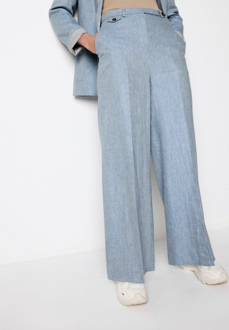 Pantaloni largi de culoare bleu deschis dintr-un material moale, având două buzunare și detalii cu nasturi la talie, asortați cu adidași albi.