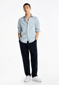Gestreepte blauw-witte shirt, met knopen, opgerolde mouwen, gecombineerd met marineblauwe broeken en witte sneakers, staand met handen in de zakken.
