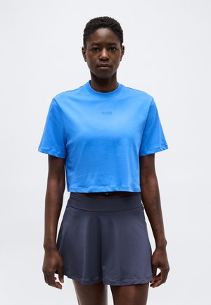 Kvinde med kort hår iført en lyseblå cropped T-shirt og en mørkegrå sportsnederdel, stående foran en ensfarvet hvid baggrund.