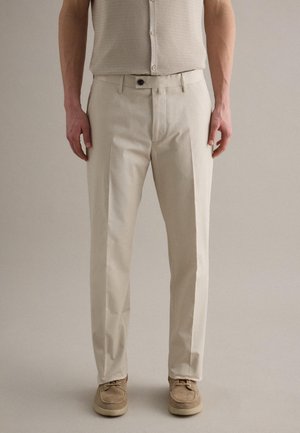 Hombre con pantalones beige hechos a medida, una camiseta sin mangas abotonada de textura beige y zapatos de gamuza marrón claro, de pie contra un fondo liso.