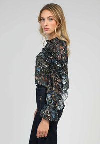 Blouse transparente à imprimé floral avec des accents à volants, fond noir, et des couleurs variées incluant du bleu et du rouge. Assortie avec un jean foncé.