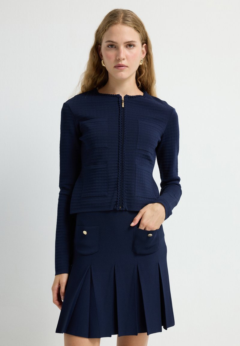 Chaqueta azul marino con textura y cremallera, adornada con detalles trenzados; combinada con una falda plisada que cuenta con dos bolsillos frontales y botones dorados.