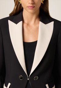 Czarna marynarka z kontrastowym kremowym kołnierzem szalowym, podwójnymi guzikami i eleganckim fasonem. Materiał wydaje się gładki z wyraźnym krojem.