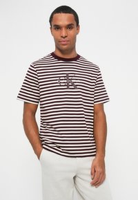 Dryžuotas trumpas rankovinis marškinėliai bordo ir grietinėlės spalvų, su apvaliu kaklu ir "Calvin Klein" logotipu ant krūtinės.