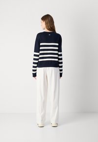 Suéter azul-marinho com riscas horizontais brancas. Mangas longas e punhos canelados, usado com calças brancas de corte largo, exibindo um design casual.