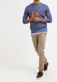 Modrá mikina s kulatým výstřihem, béžové kalhoty a hnědé tenisky. Mikina má strukturovaný povrch a žebrované manžety; kalhoty jsou úzkého střihu.