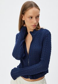 Mörkblå stickad cardigan med dragkedja framtill och ribbad krage. Den texturerade designen inkluderar kabelstickade mönster och åtsittande ärmar.