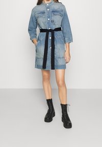 Denimklänning med krage, framknappar, två bröstfickor och ett svart bälte. Bärs med svarta snörning plattformstövlar. Ljusblå färg.