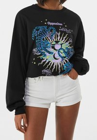 Maglietta nera a maniche lunghe con grafica colorata di una tigre e del sole, con la parola "Opposites" sopra, indossata con pantaloni corti bianchi a orlo arrotolato, mano in tasca.