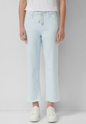 Person trägt hellblaue Jeans mit geradem Bein und ausgefransten Säumen, weiße Turnschuhe und ein weißes Hemd vor einem einfarbigen Hintergrund.