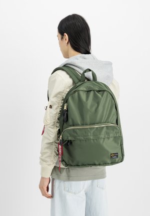 Mochila de nylon verde com um design elegante, apresentando um bolso frontal com fecho zip, alças duplas para transporte, e etiquetas vermelhas. Usada com um moletom cinza claro.