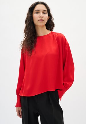 Mujer con cabello largo y rizado, vestida con una blusa roja holgada y pantalones negros, de pie con una mano en el bolsillo frente a un fondo blanco liso.