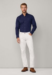 Marineblauw overhemd met knopen en een klein logo, gecombineerd met witte jeans en bruine veterschoenen, staande tegen een neutrale achtergrond.