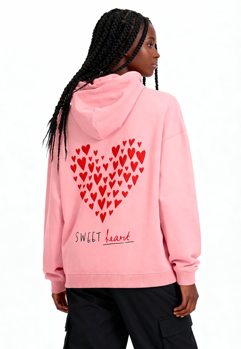 Felpa rosa con un grande design a forma di cuore composto da cuori rossi sul retro, con il testo "SWEET heart" in nero e polsini a coste.