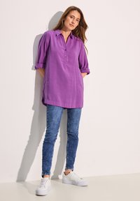Camicia tunica viola con pattina buttonata, maniche arrotolate, abbinata a jeans skinny blu e sneaker platform bianche su uno sfondo chiaro.