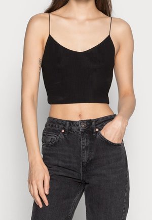 Frau trägt ein schwarzes geripptes Croptop mit dünnen Trägern und hoch taillierte ausgebleichte schwarze Jeans, eine Hand in der Tasche, vor einem schlichten Hintergrund.