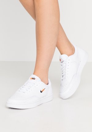 Białe sneakersy Nike z czarnym i pomarańczowym logo noszone na gołych stopach, nogi skrzyżowane na tle jednolitego jasnego tła.