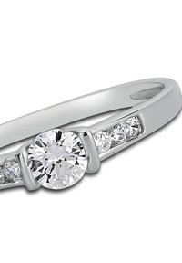 Bague en métal blanc avec un diamant rond central entouré de diamants plus petits de chaque côté, arborant un design élégant et poli.