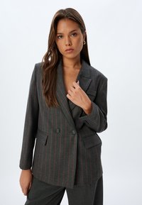 Blazer gris à double boutonnage avec de subtils rayures rouges. Présente des revers crantés, deux poches frontales et des fermetures à boutons. Tissu texturé.