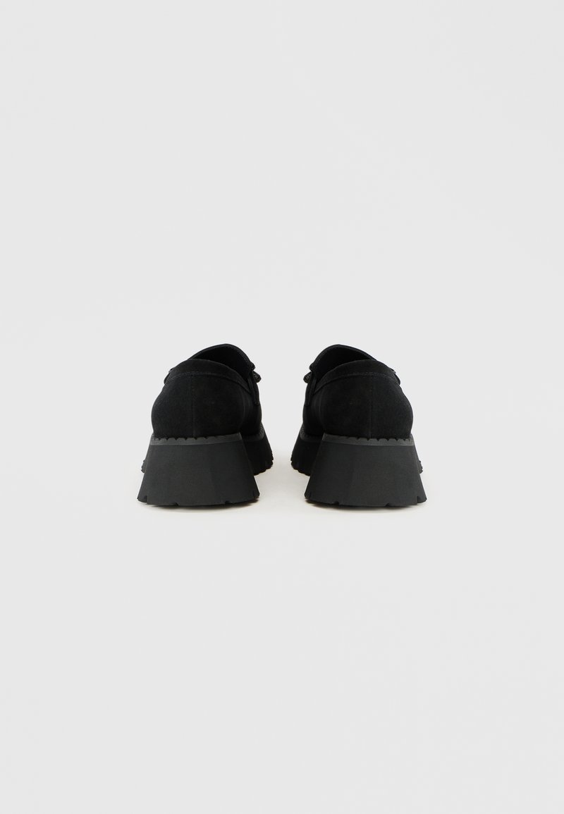 Czarne zamszowe buty na grubej, chropowatej czarnej gumowej podeszwie, o zaokrąglonym kształcie i minimalistycznym wzornictwie, widziane z tyłu.