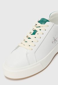 Bijele tenisice Calvin Klein s kremastim vezicama, zelenim jezikom i logotipom sa strane, s debljim bijelim đonom i minimalističkim dizajnom.