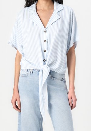 Femme portant un chemisier à rayures verticales bleu clair et blanc, boutonné et noué à la taille, associé à un jean en denim bleu clair.