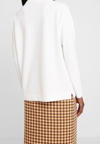 Maglione bianco a trama con maniche lunghe, caratterizzato da spacchi laterali, abbinato a una gonna con motivo pied de poule marrone e giallo.