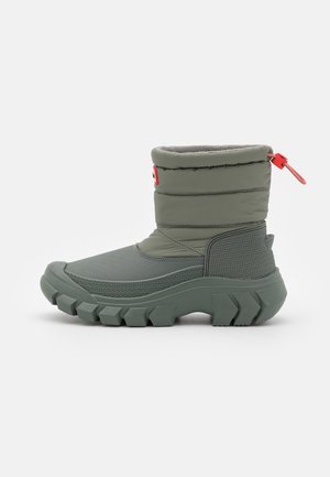 Botte d’hiver verte avec semelle épaisse et texturée, partie rembourrée à la cheville et fermeture à bascule rouge à l’arrière.